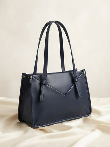 Knot-accent structured shoulder bag in navy with top handles, diagonal seam, and gold studs